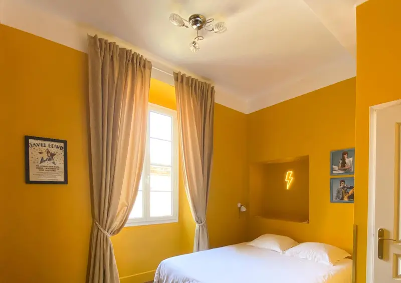 Une chambre lumineuse aux murs jaunes est décorée de rideaux beiges et d'œuvres d'art.