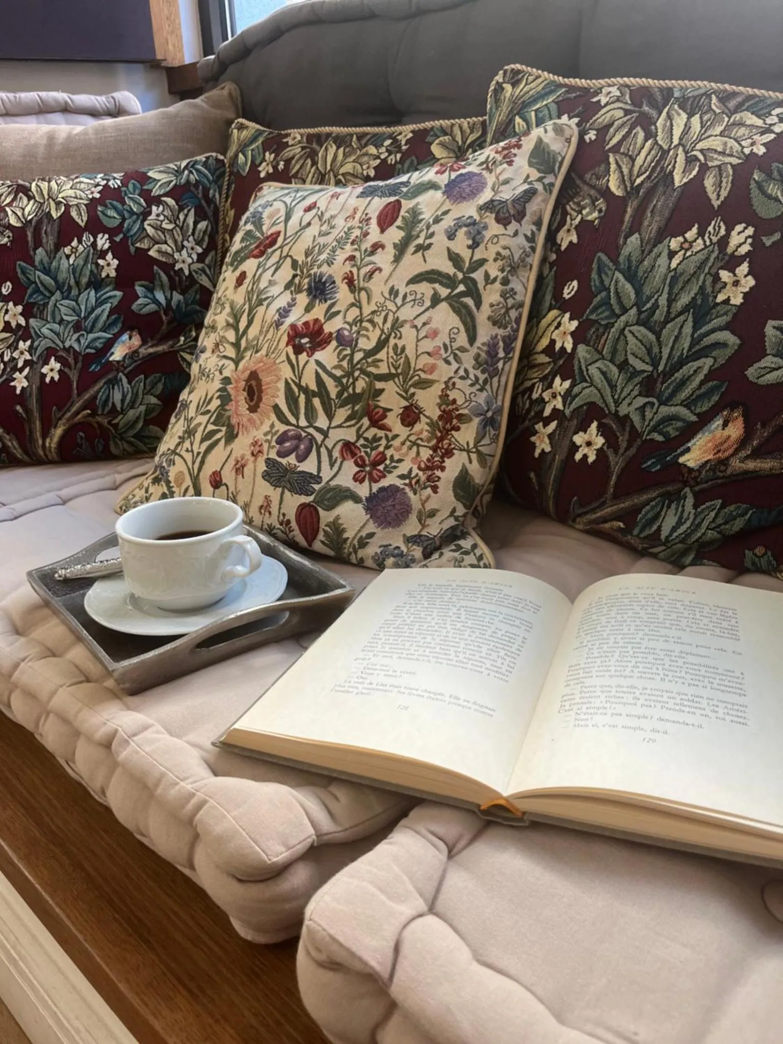 Un livre ouvert et une tasse de café reposent sur un canapé agrémenté de coussins fleuris.