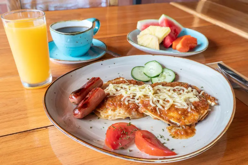 Une assiette copieuse avec des œufs au fromage, des saucisses, des concombres, des tomates et des fruits frais.