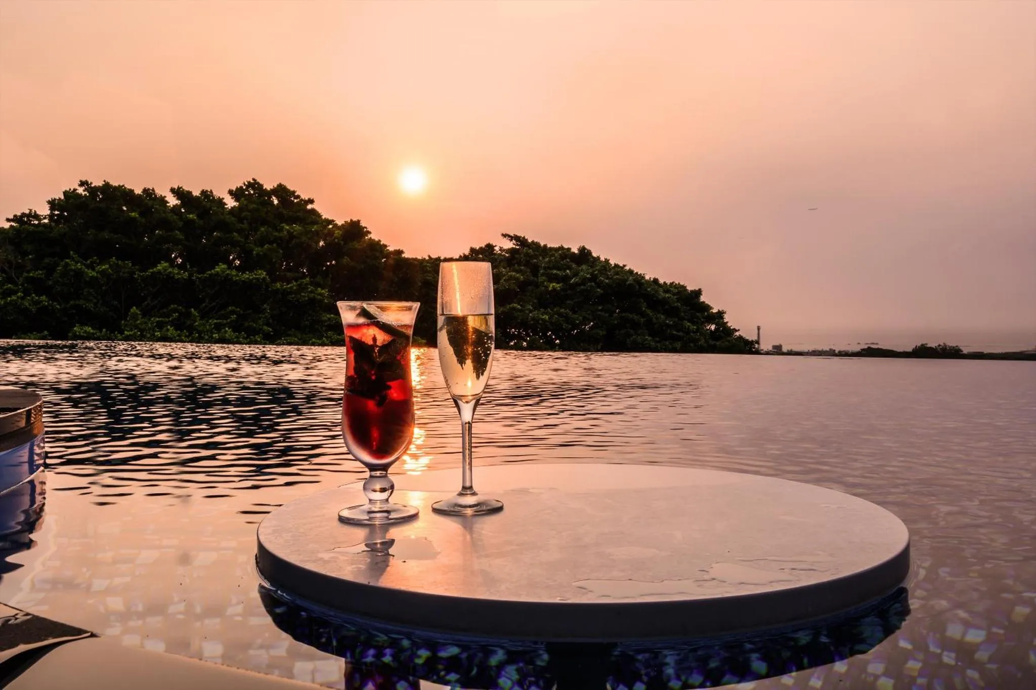 Deux verres colorés reposent sur une table au bord d'une piscine, avec un coucher de soleil en arrière-plan.