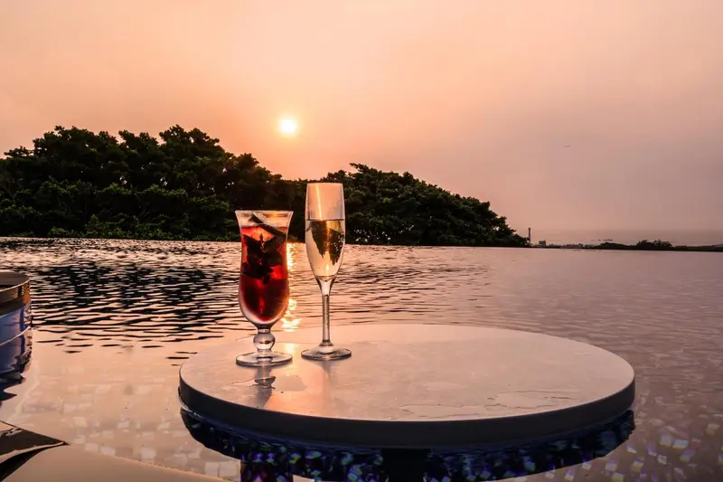 Deux verres colorés reposent sur une table au bord d'une piscine, avec un coucher de soleil en arrière-plan.