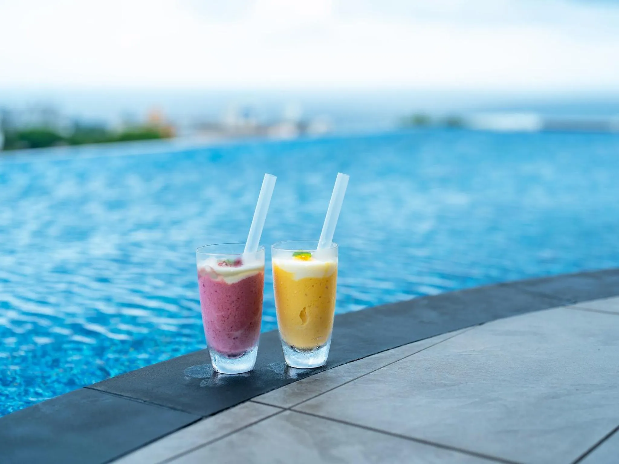 Deux cocktails colorés reposent sur le bord d'une piscine bleu clair face à la mer.