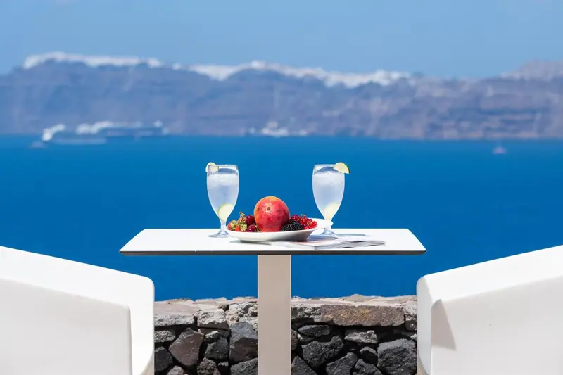 Deux verres et une assiette de fruits sont posés sur une table blanche face à la mer.