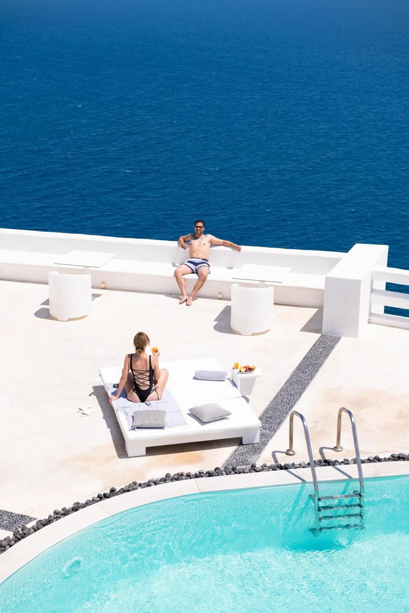 Deux personnes se détendent sur des transats blancs au bord d'une piscine à débordement avec vue sur la mer.
