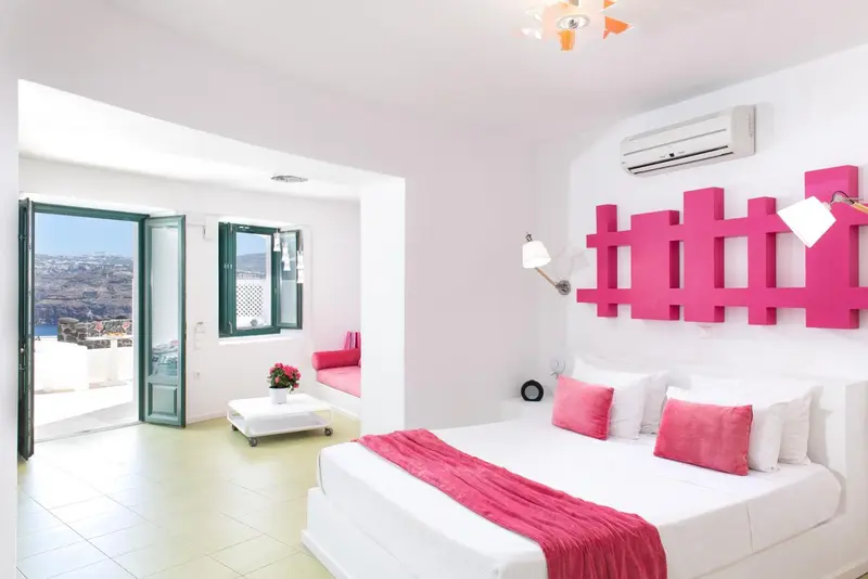 Chambre lumineuse avec un lit blanc et des touches de rose, donnant sur une vue sur la mer.