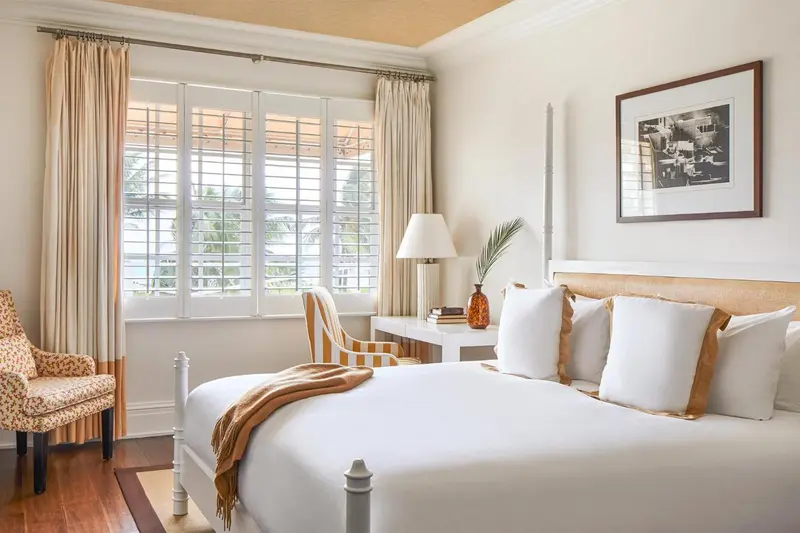Une chambre lumineuse avec un lit recouvert d'une courtepointe blanche et des rideaux beiges.