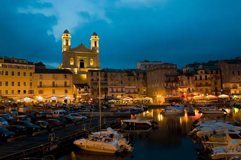 Vue nocturne d'un port de plaisance avec une église illuminée en arrière-plan.