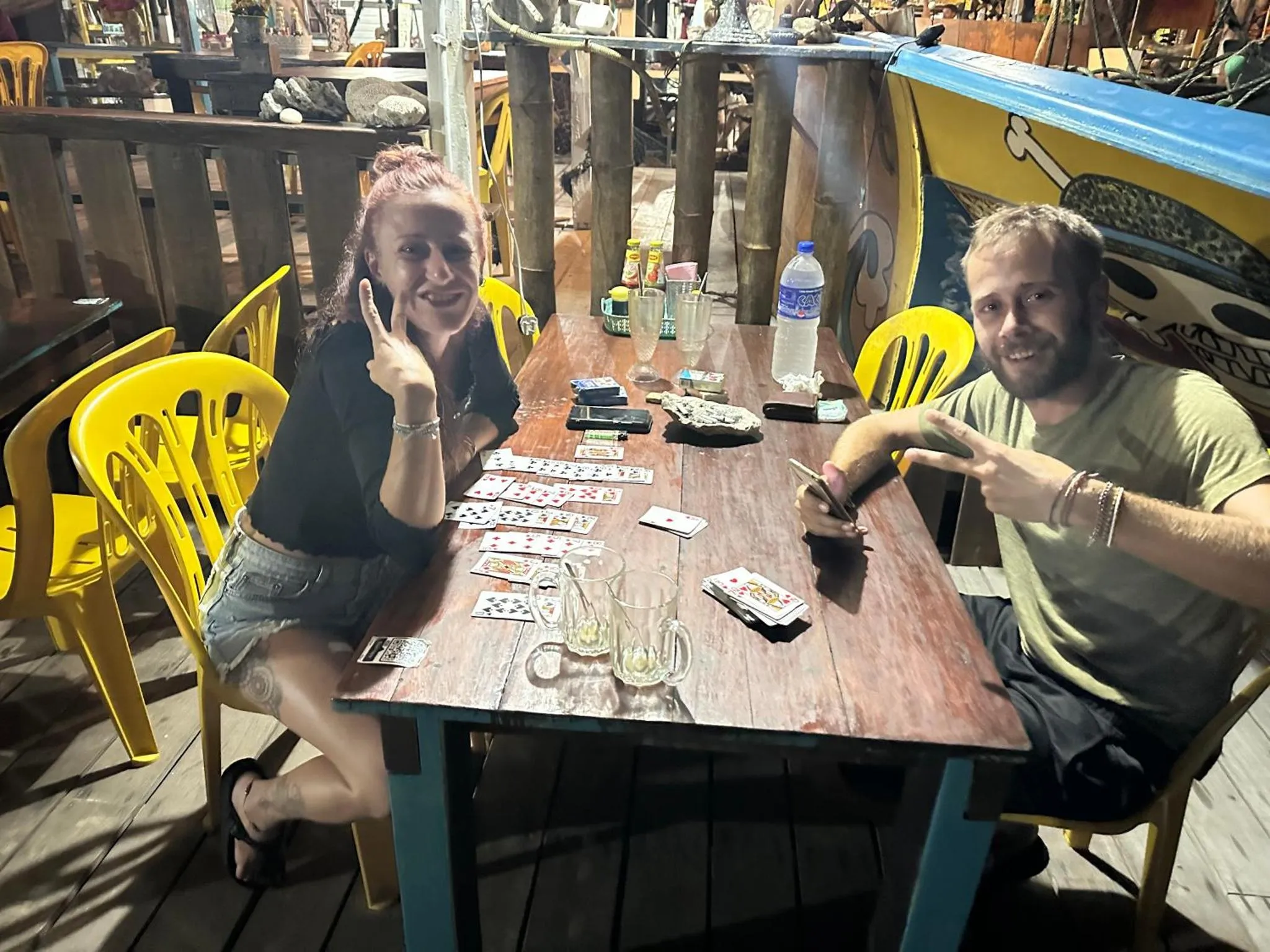 Deux personnes sourient en jouant aux cartes à une table en bois sur une terrasse.