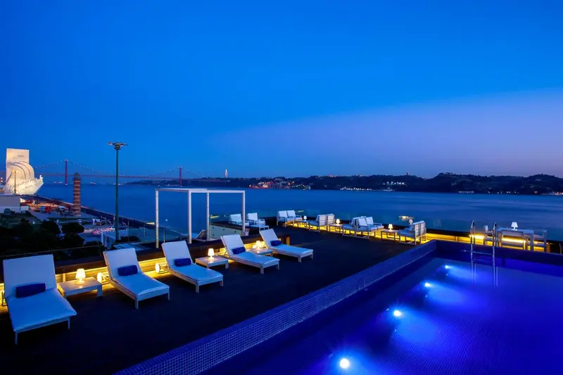 Terrasse avec transats et piscine au bord de l'eau, éclairée la nuit.