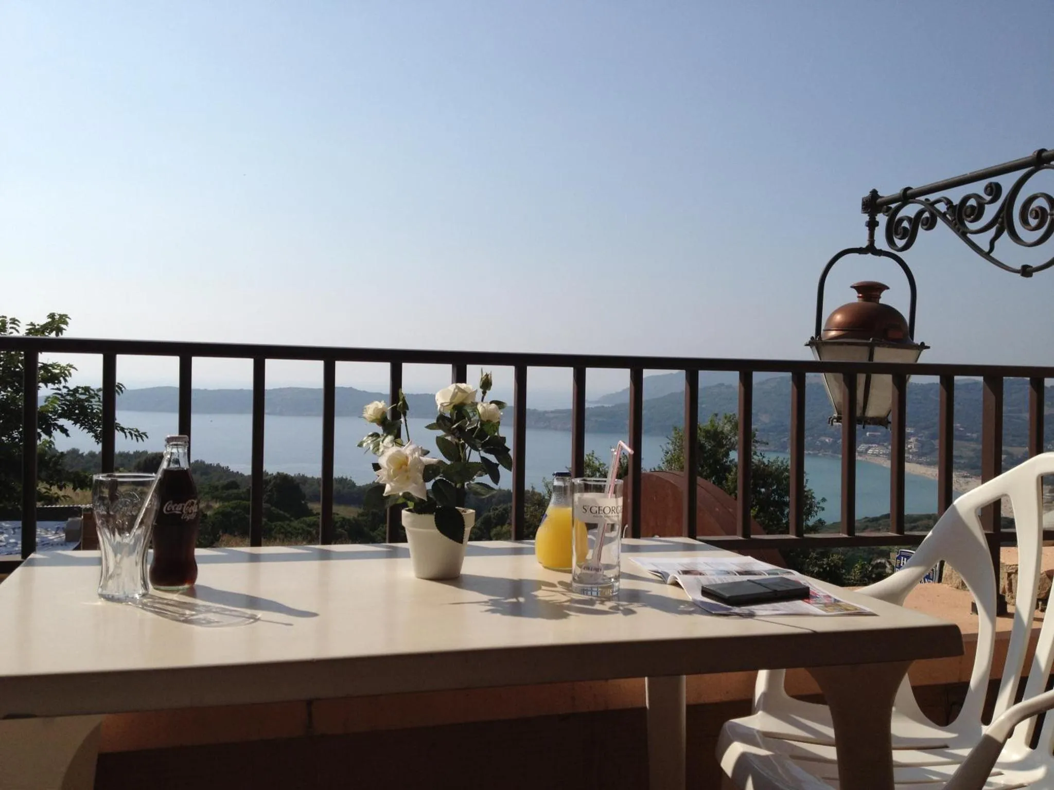 Une table avec des boissons et un journal est placée sur une terrasse ensoleillée avec vue sur la mer.