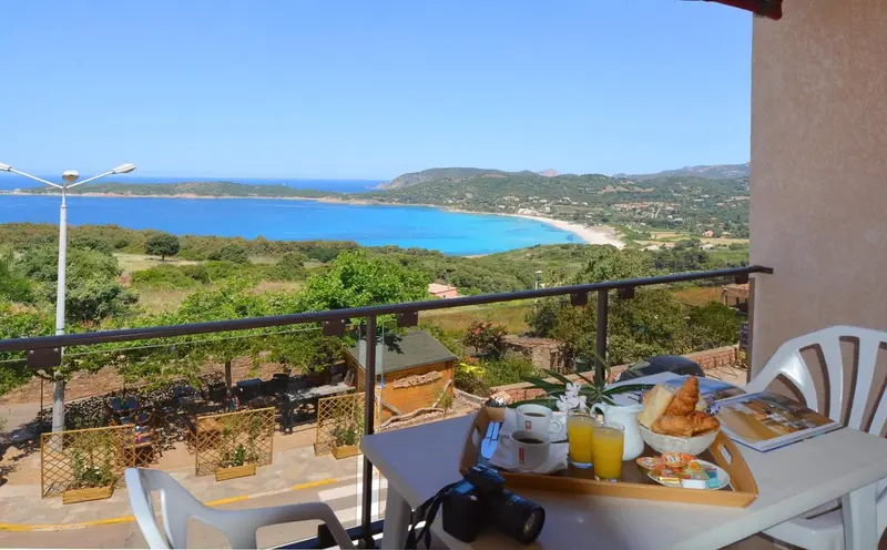 Une table de petit-déjeuner est dressée sur une terrasse avec vue sur la mer et les collines.