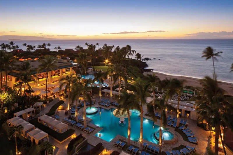 Vue aérienne d'un complexe hôtelier illuminé au coucher du soleil, avec des piscines et des palmiers.