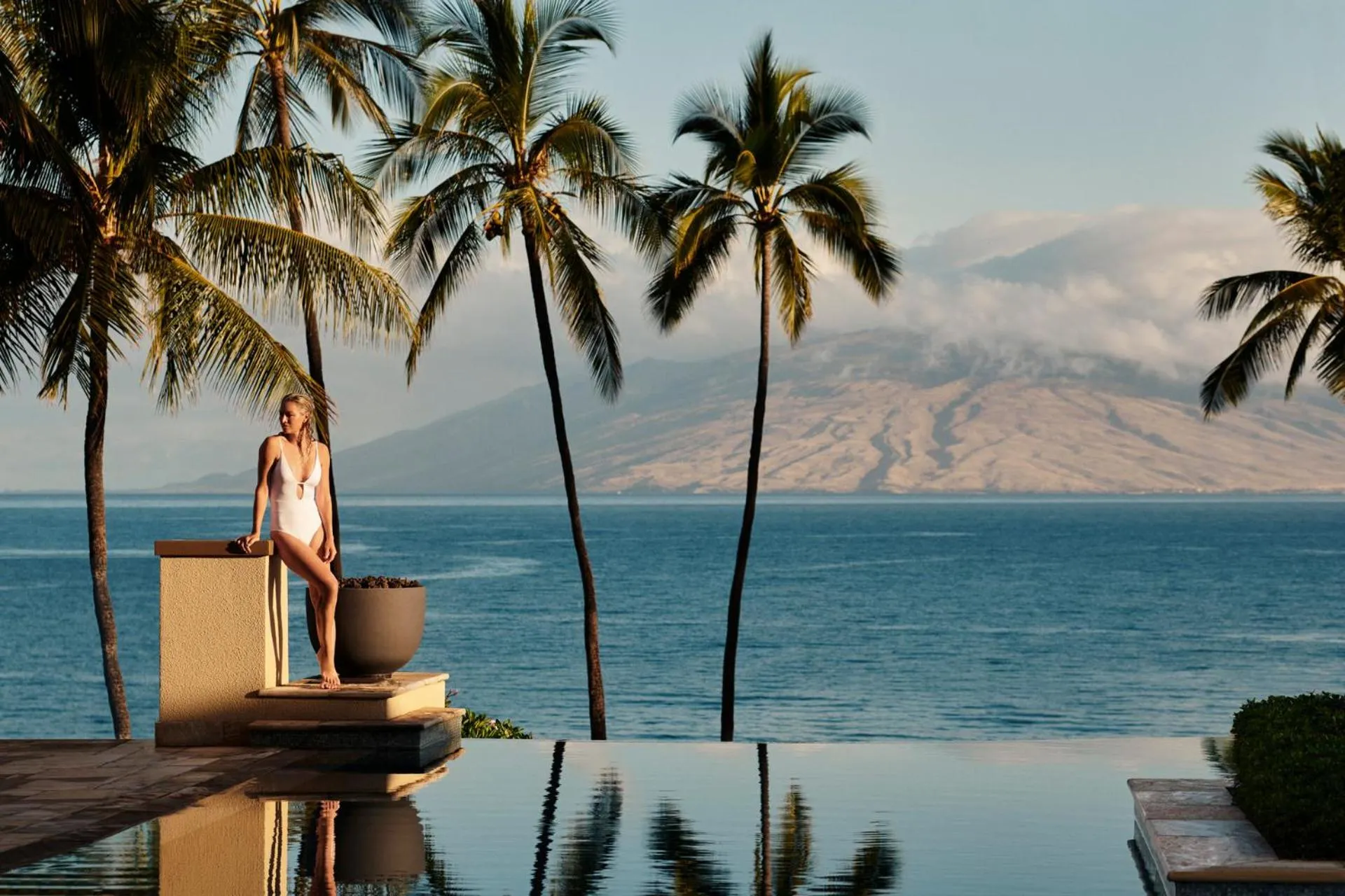 Une femme se tient près d'une piscine à débordement avec vue sur la mer et les montagnes.