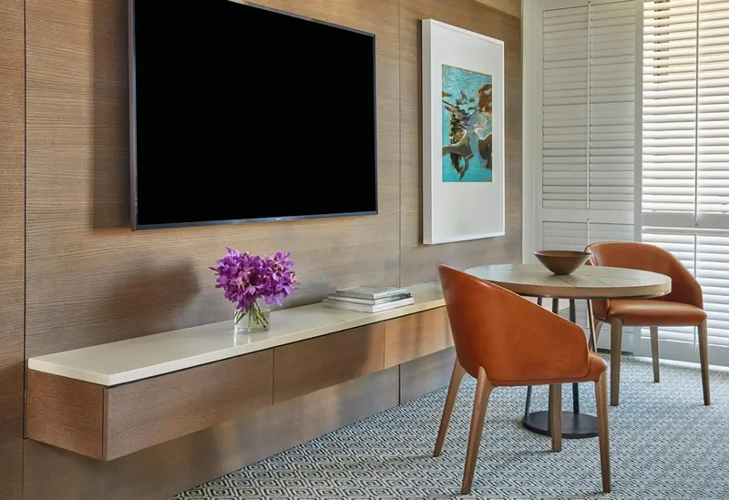 Une télévision murale domine un meuble bas avec un bouquet de fleurs et une table ronde avec deux chaises.