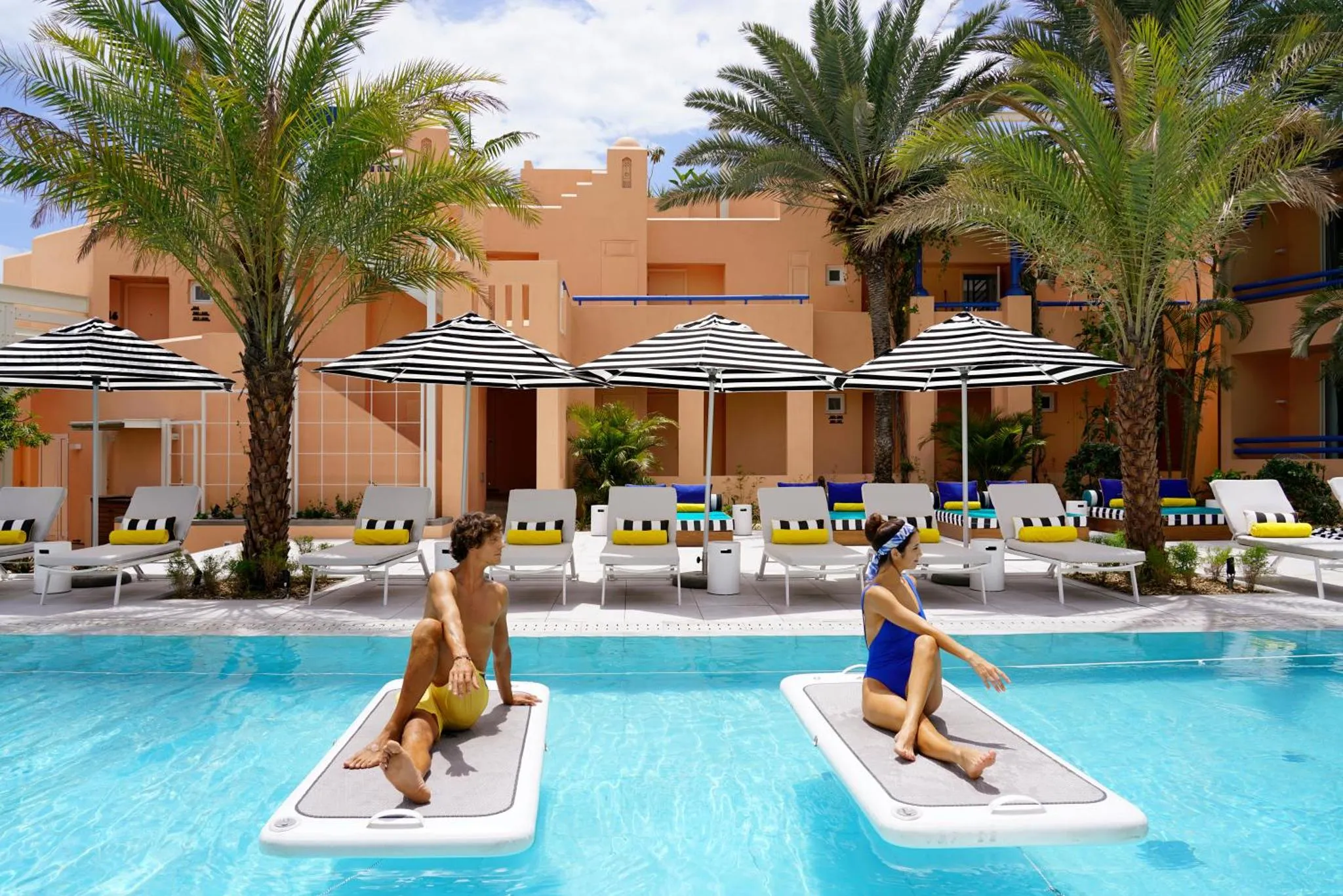 Deux personnes assises sur des planches flottantes dans une piscine ensoleillée, entourée de transats et de parasols.