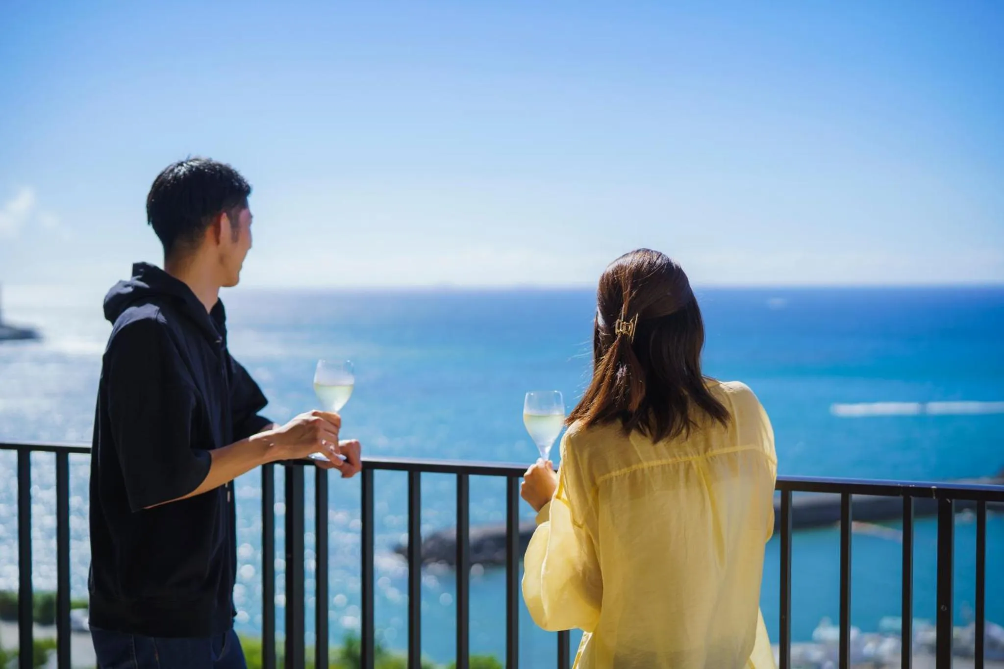 Un couple se tient sur un balcon avec des verres à la main, face à la mer.