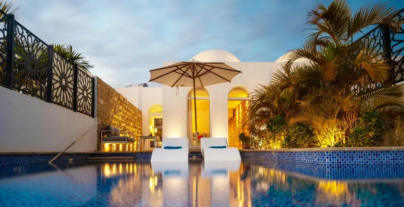 Deux fauteuils blancs sont placés au bord d'une piscine illuminée, devant une façade blanche ornée d'arches.