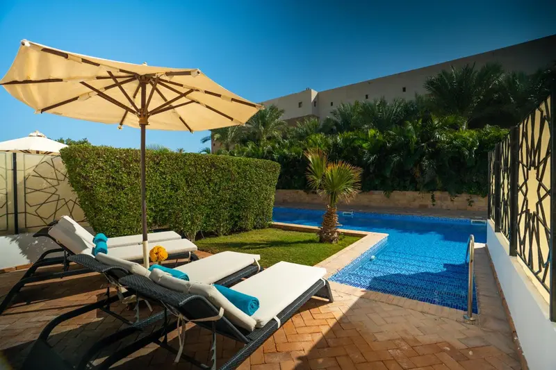 Transats et parasol ombragent une piscine rectangulaire dans un jardin soigné.