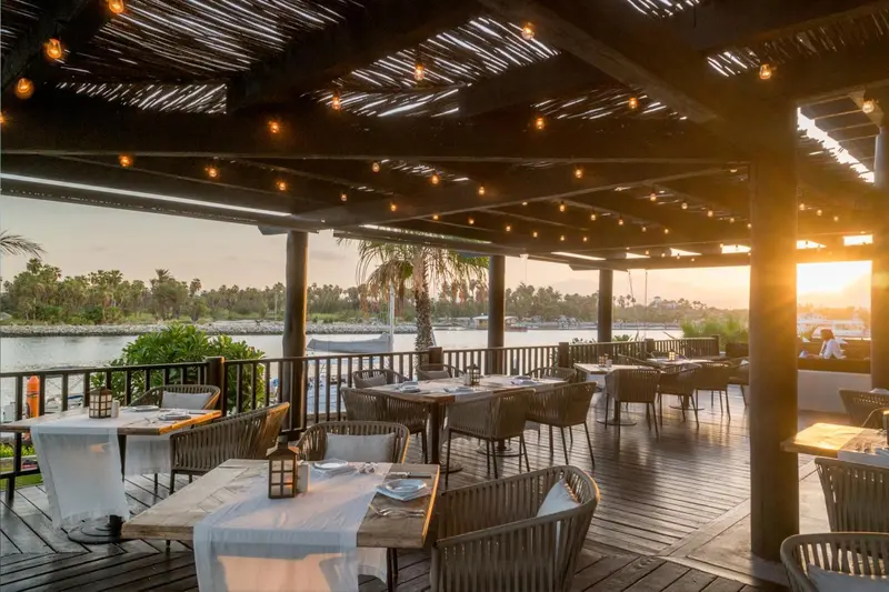 Terrasse ombragée avec tables dressées face à un plan d'eau au coucher du soleil.