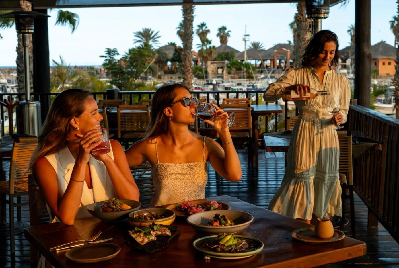 Trois femmes sont assises à une table en bois sur une terrasse, profitant de boissons et de repas.