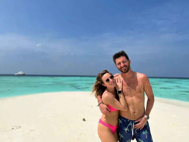 Un couple souriant se tient sur une plage de sable blanc face à une mer turquoise.