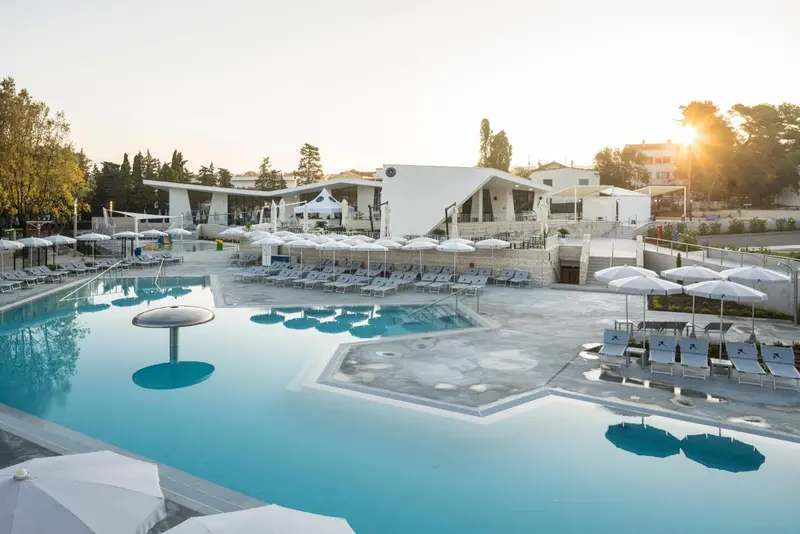 Piscine turquoise avec transats blancs et parasols sous le soleil couchant.