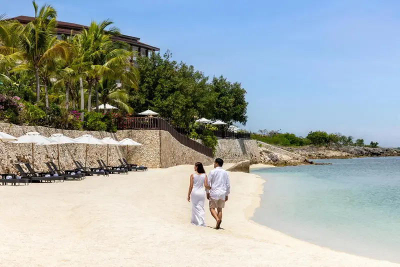 Un couple se promène sur une plage de sable blanc bordée de palmiers.