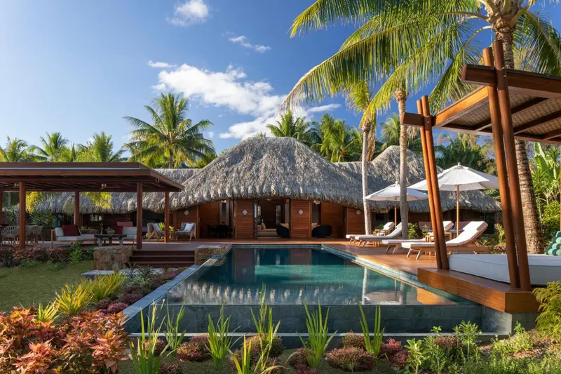 Une piscine turquoise bordée de chaises longues devant des bungalows aux toits de chaume.
