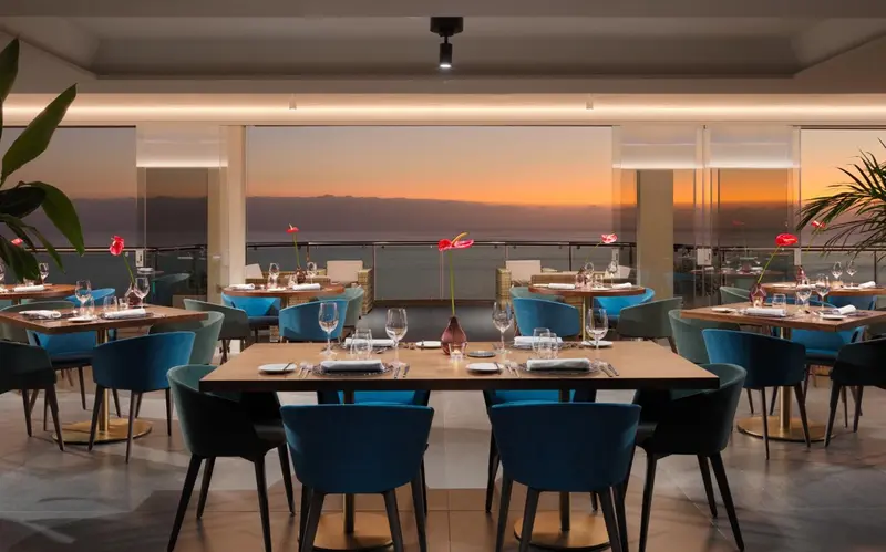 Une salle à manger lumineuse avec des tables dressées face à une vue sur la mer au coucher du soleil.
