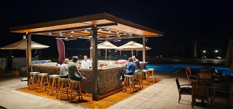 Un bar extérieur illuminé avec des clients assis au comptoir et sur des tabourets.