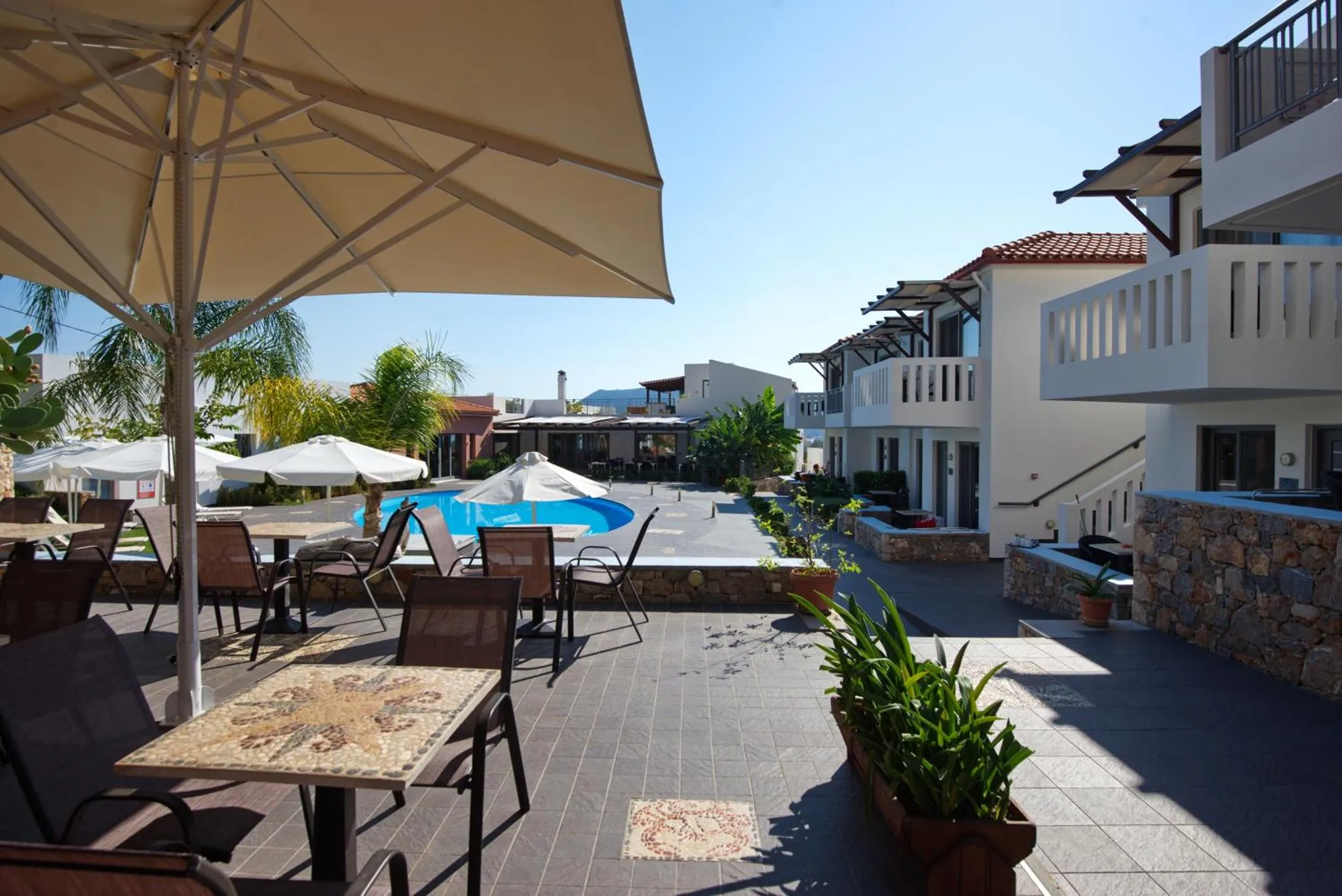 Terrasse ombragée avec tables et chaises donnant sur une piscine et des bâtiments blancs.