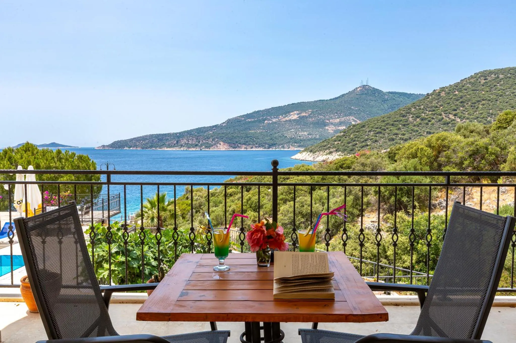 Une table avec des boissons et un livre est placée sur une terrasse avec vue sur la mer et les collines.