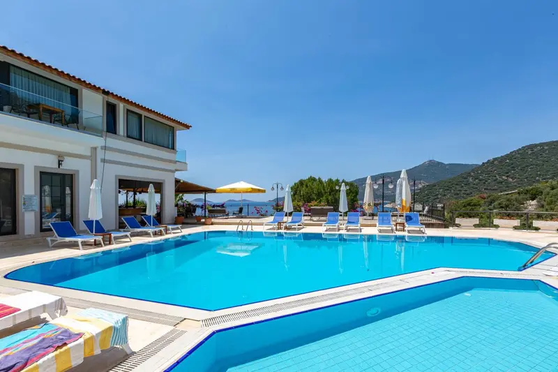Piscine bleue bordée de transats devant un bâtiment blanc et une colline verdoyante.