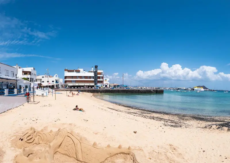 Plage de sable fin avec des personnes se relaxant au soleil et des bateaux amarrés au loin.