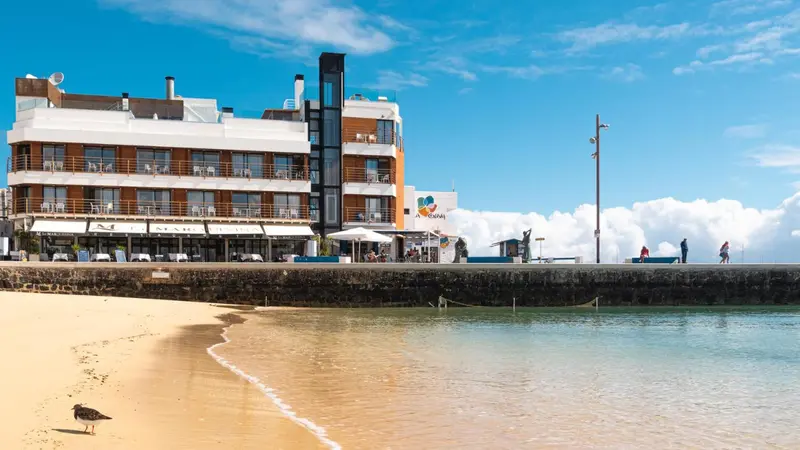 Bâtiment blanc et beige avec des balcons donnant sur une plage de sable et une mer bleue.