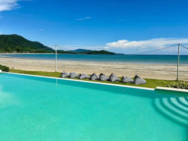 Une piscine turquoise bordée de chaises longues face à une plage de sable fin et une mer bleue.
