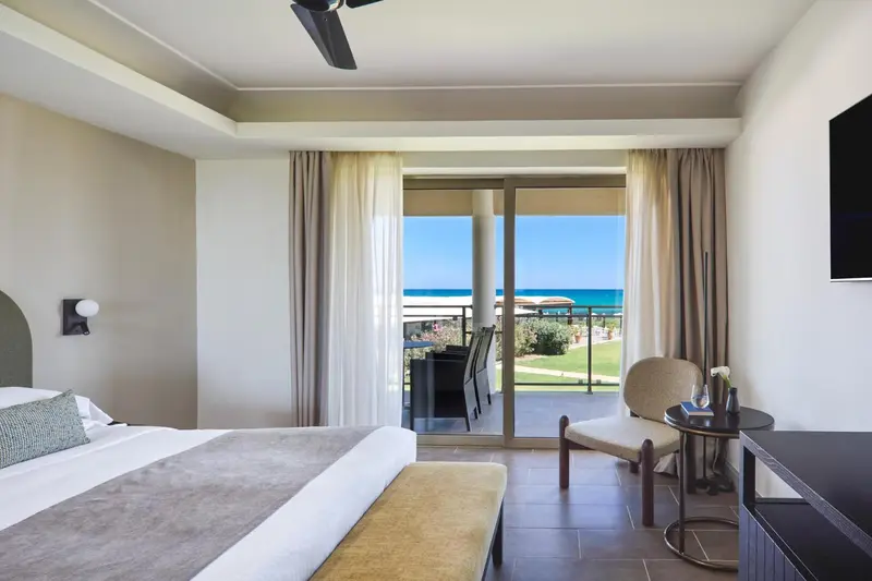 Une chambre lumineuse avec un lit fait et un fauteuil près d'une baie vitrée donnant sur la mer.