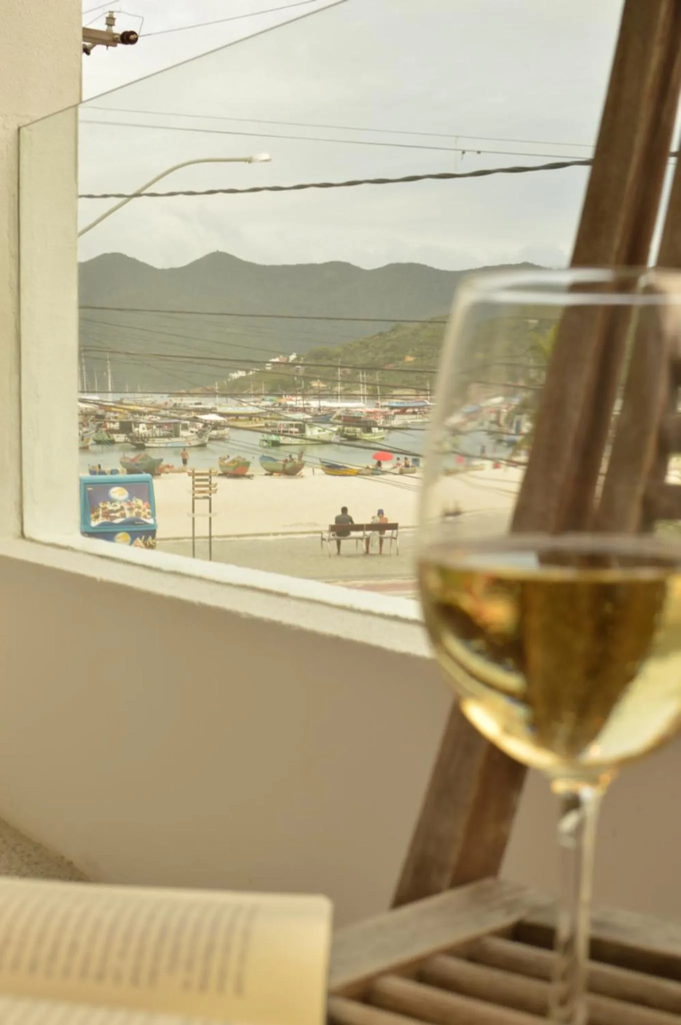 Vue sur une plage et des montagnes depuis une fenêtre avec un verre de vin au premier plan.