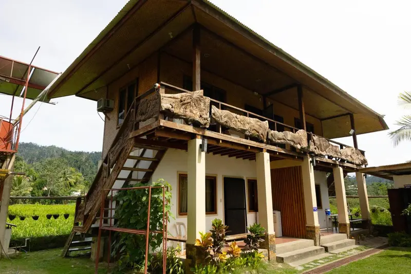 Bâtiment en bois sur pilotis avec balcon orné et végétation luxuriante.