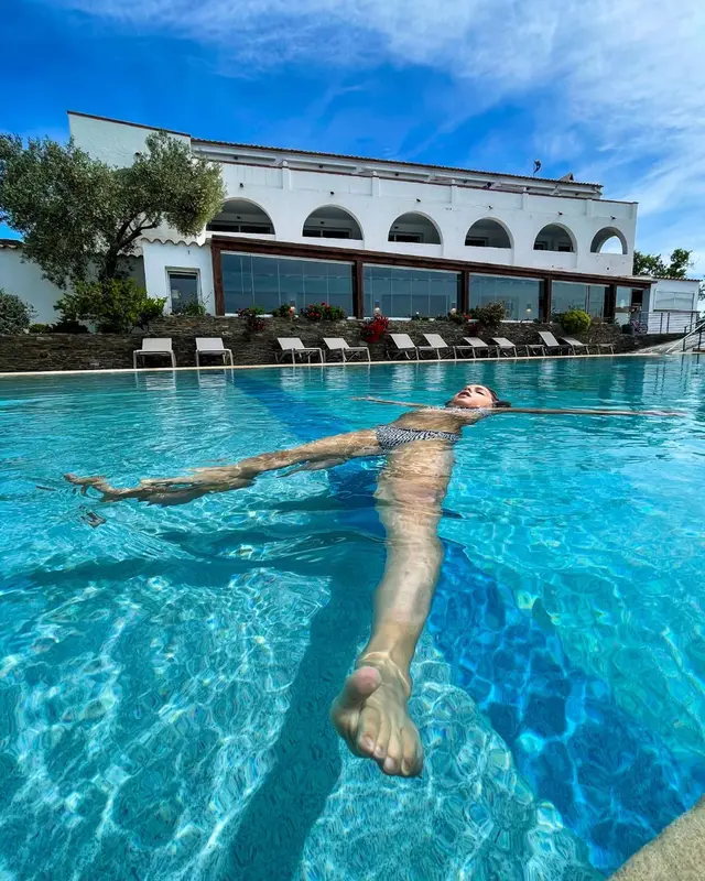 Une personne flotte sur le dos dans une piscine ensoleillée devant un bâtiment blanc.