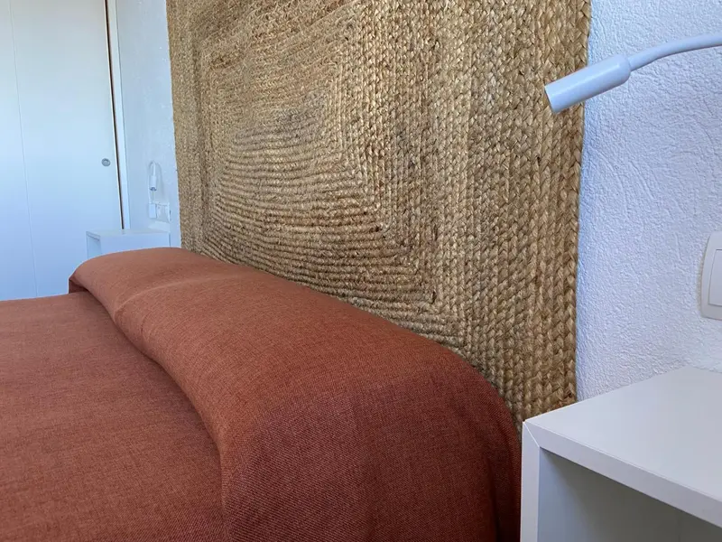 Un lit recouvert d'une couverture terracotta est adossé à une tête de lit en fibres naturelles.