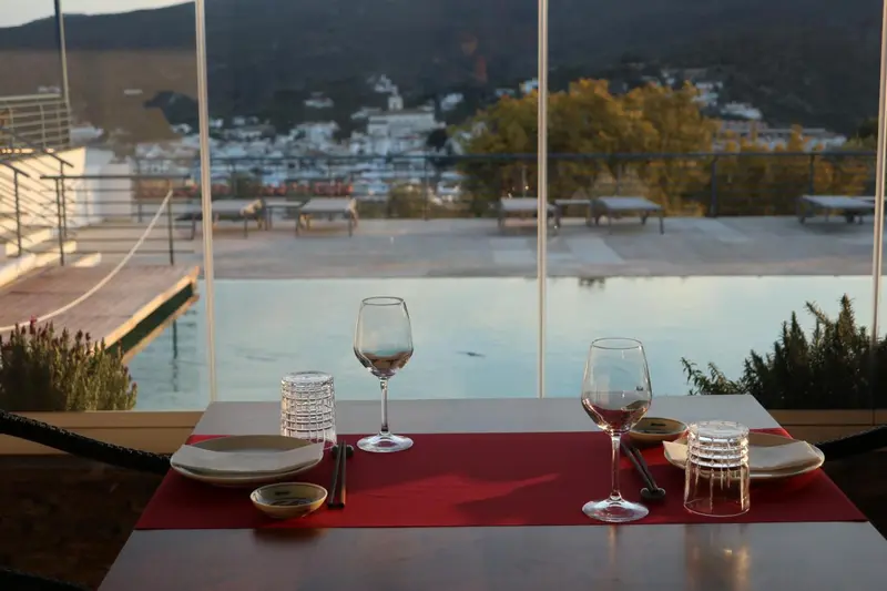 Une table dressée avec de la vaisselle et des verres, devant une grande baie vitrée donnant sur une piscine et un paysage urbain.