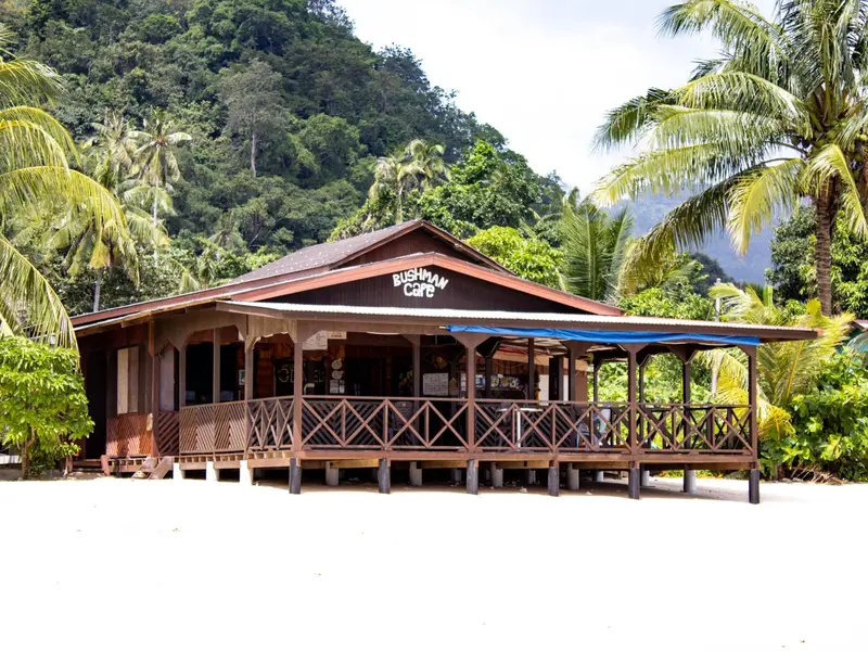 Bâtiment en bois sur pilotis face à une plage de sable blanc bordée de végétation tropicale.