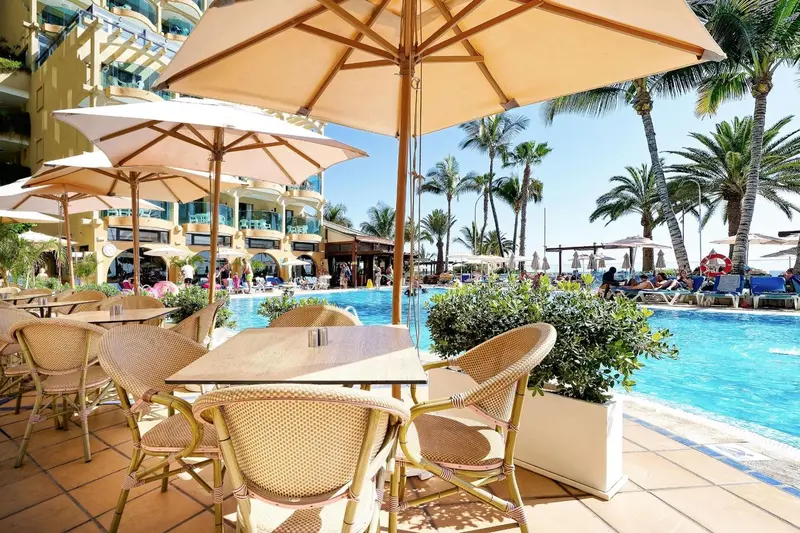 Tables et chaises en osier disposées près d'une piscine ensoleillée avec des palmiers en arrière-plan.