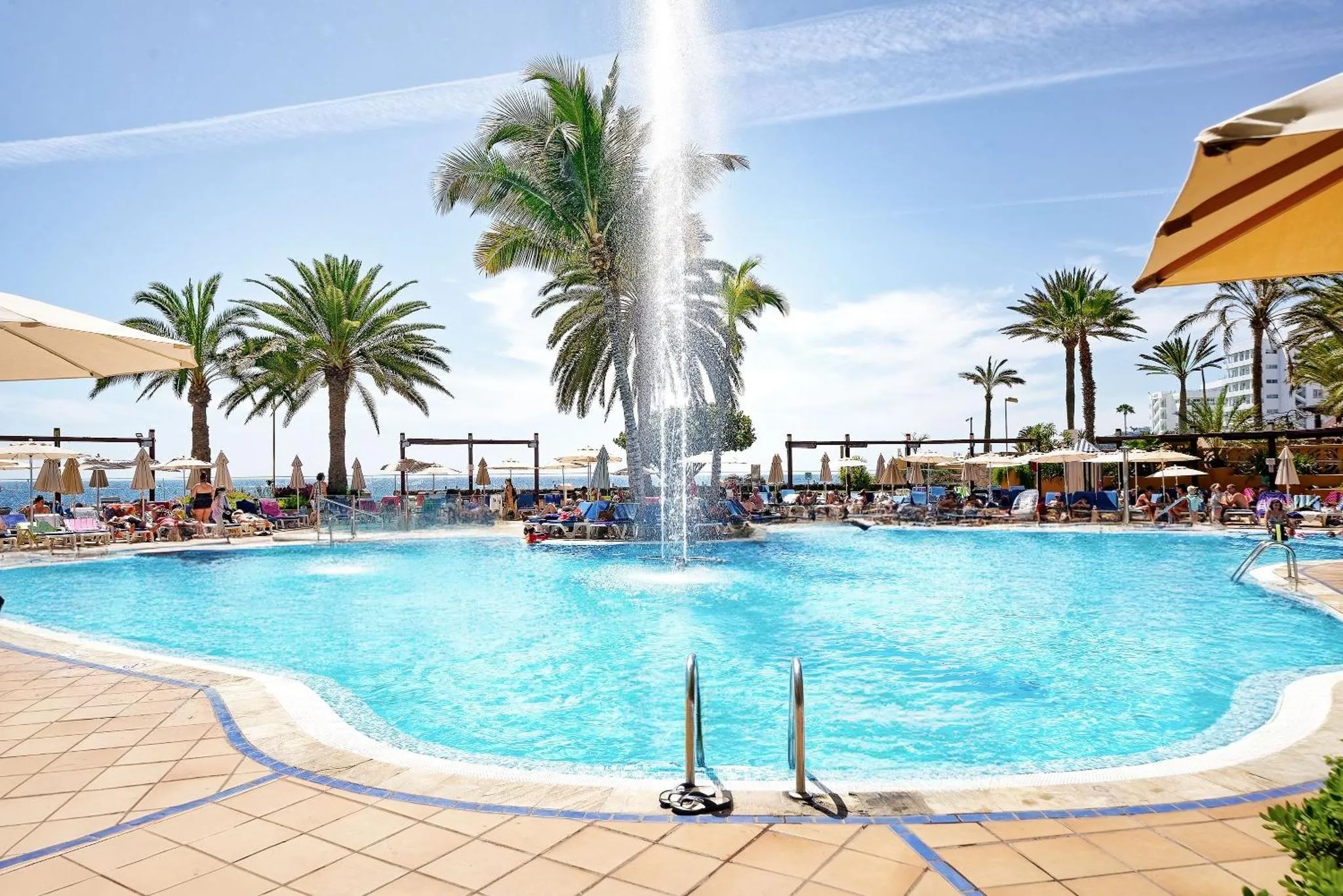 Une piscine turquoise entourée de transats et de palmiers sous le soleil.