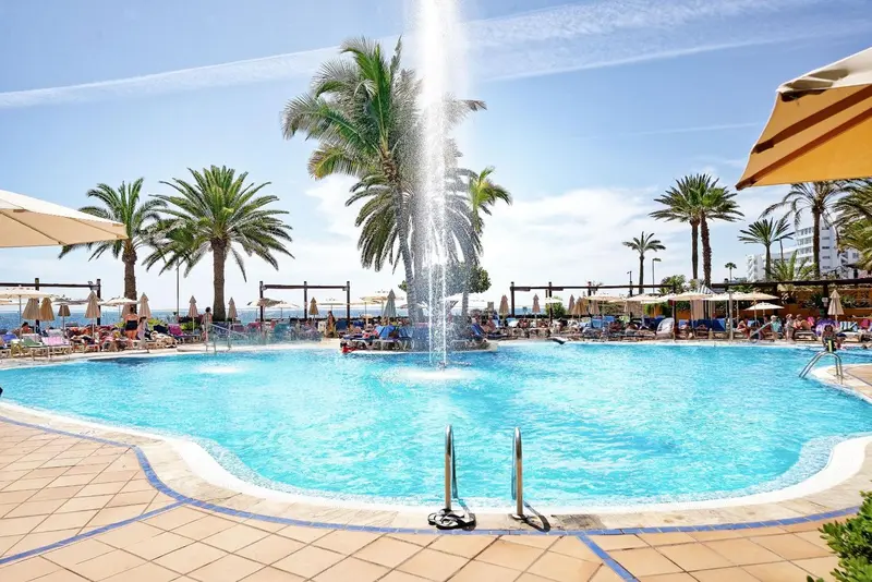Une piscine turquoise entourée de transats et de palmiers sous le soleil.