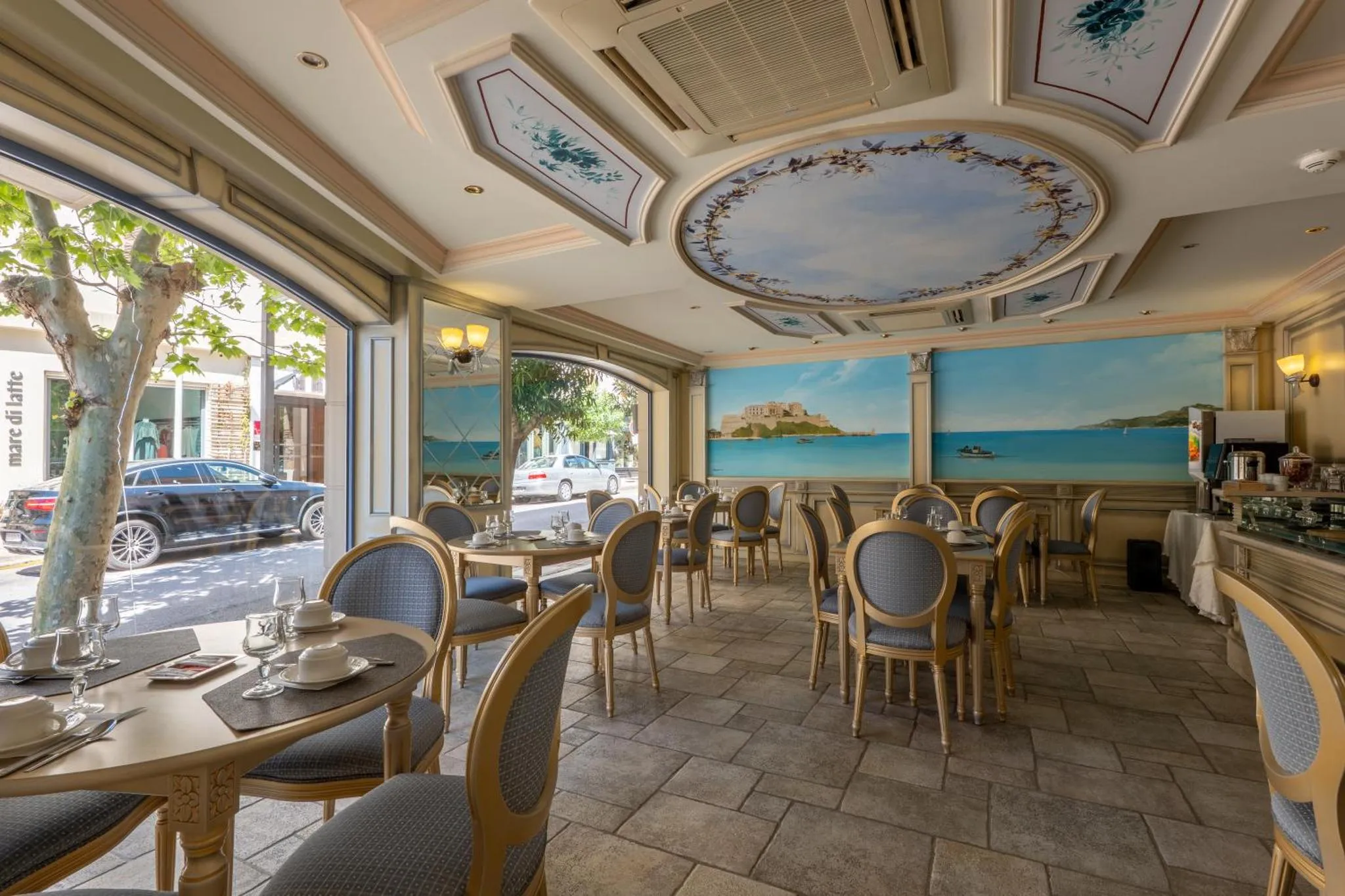 Une salle lumineuse avec des tables dressées devant une grande fenêtre donnant sur une rue et la mer.