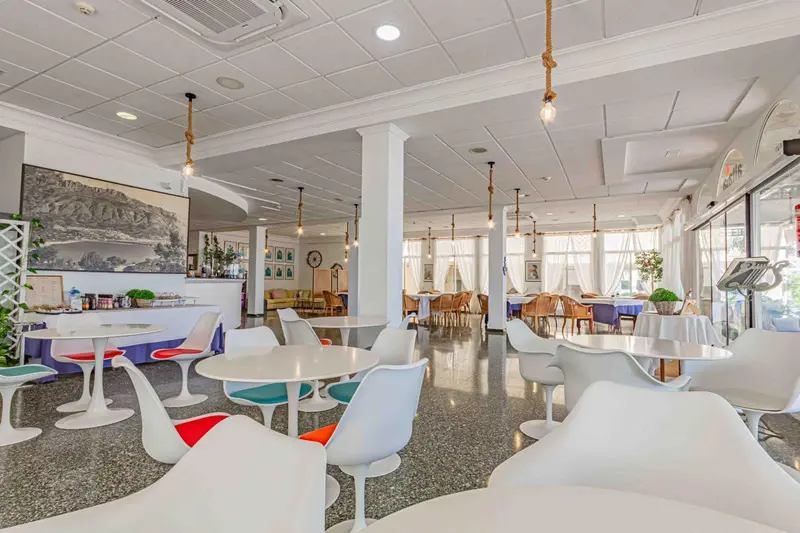 Une salle lumineuse avec des tables et des chaises blanches, décorée de plantes et d'éclairages suspendus.