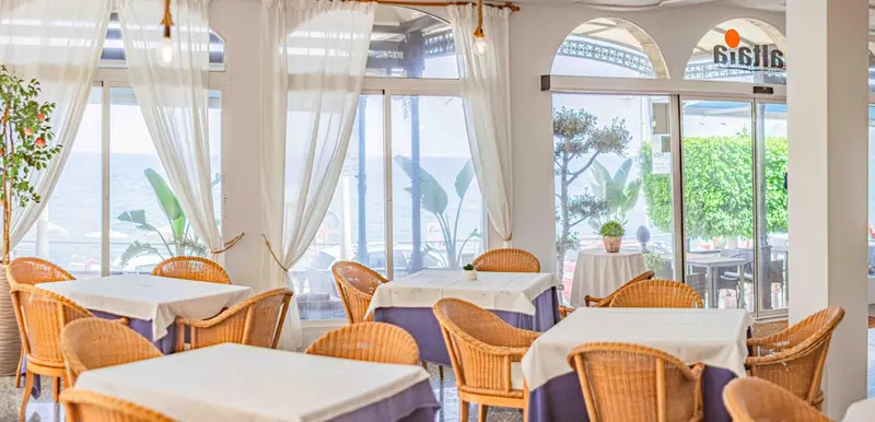 Une salle à manger lumineuse avec des tables nappées et des chaises en osier, donnant sur la mer.