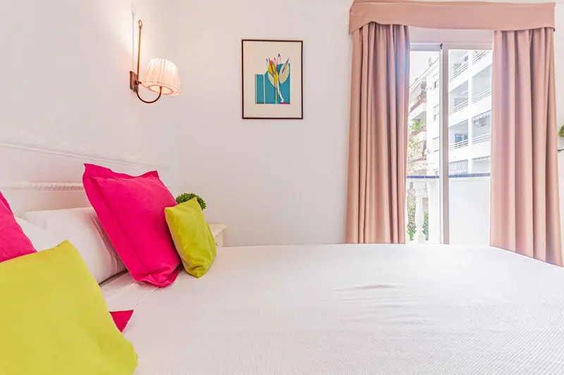 Lit blanc avec coussins roses et verts dans une chambre lumineuse.